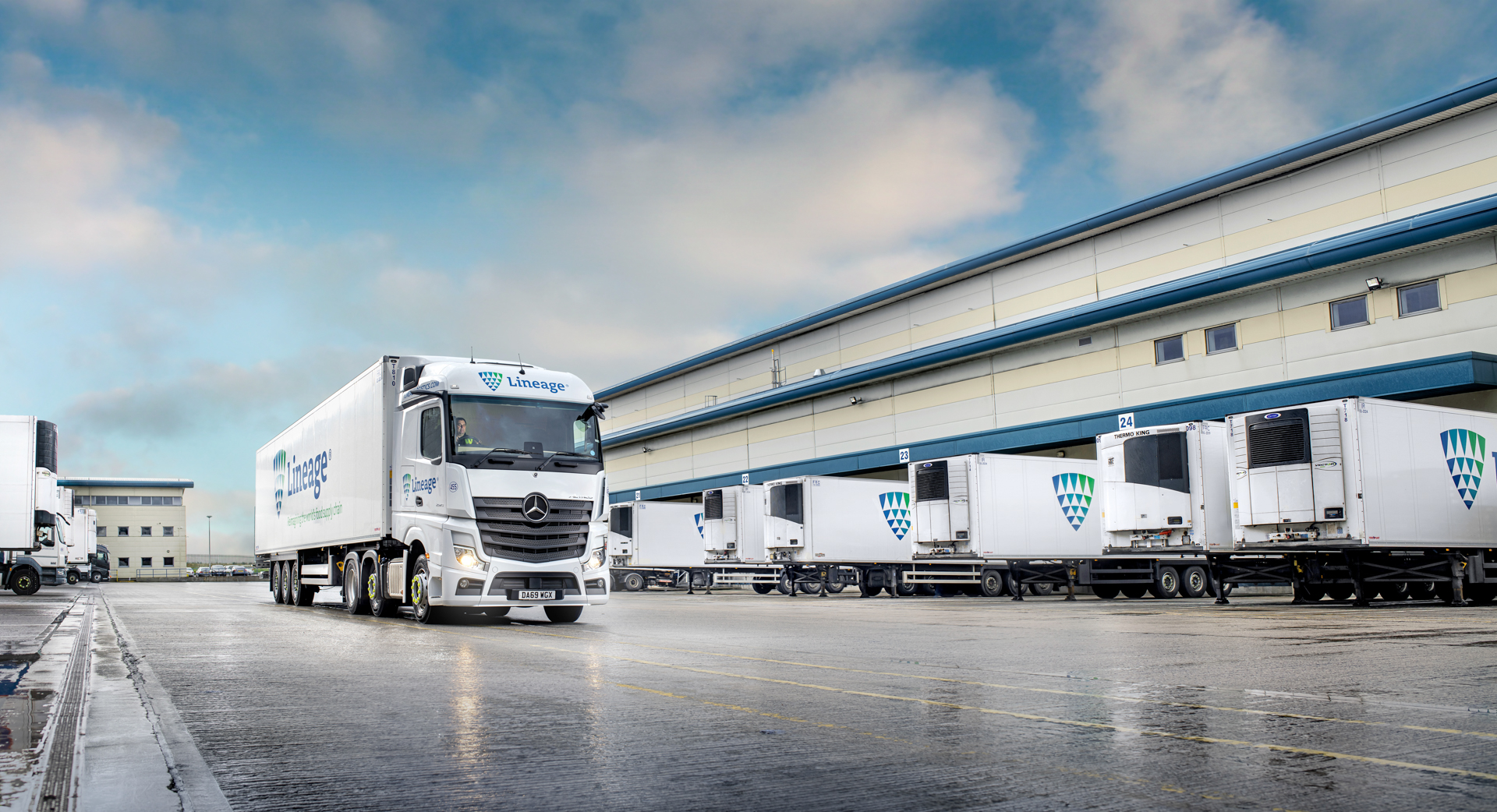 A Lineage truck driving past a fleet of parked Lineage trucks outside a Lineage warehouse.