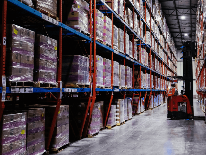 Interior view of a cold storage warehouse with high shelves packed with pallets wrapped in purple plastic, under dim lighting. A forklift is operated by a worker in the central aisle.